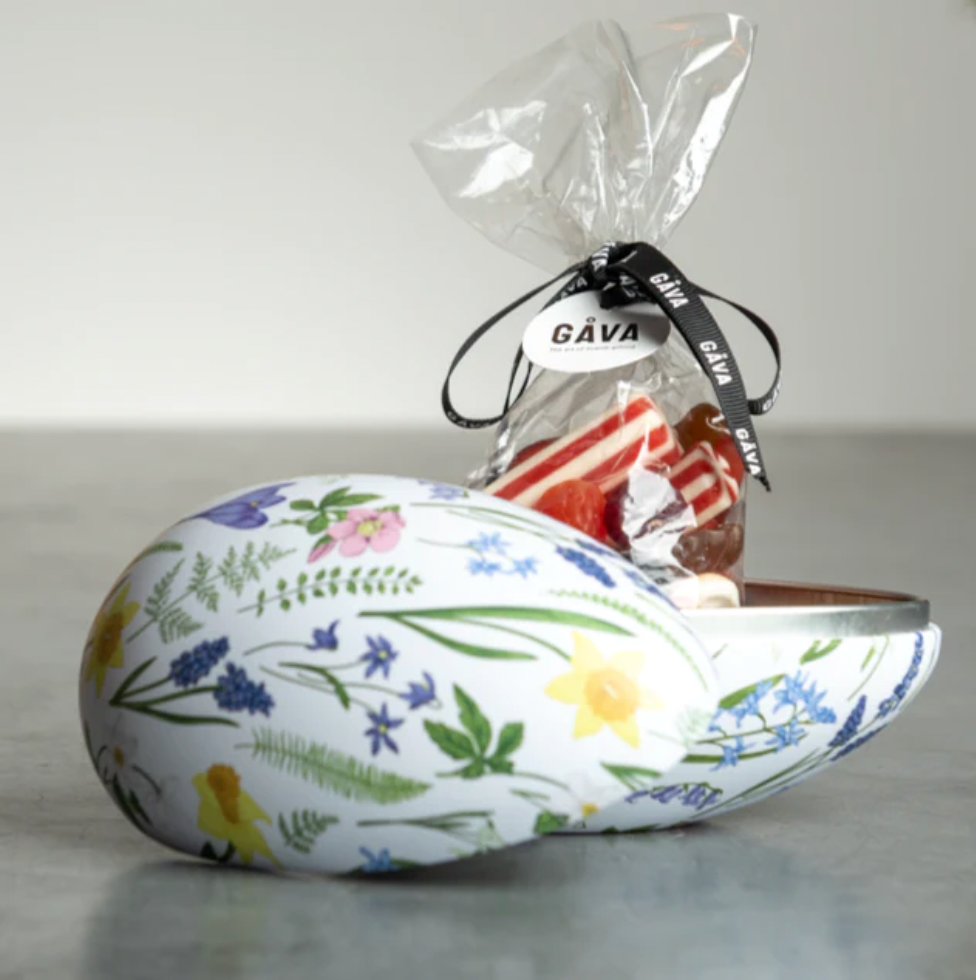 A decorative egg-shaped container with a floral pattern, partially open to reveal a bag of candy tied with a black ribbon labeled GÅVA.