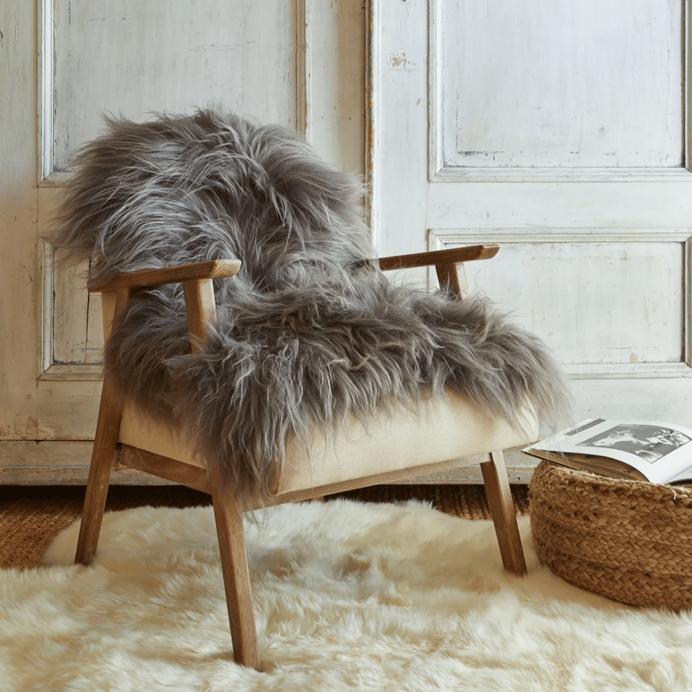 A cozy wooden armchair with a gray faux fur throw draped over it sits on a white fluffy rug next to a woven basket and an open book, in front of rustic white paneled doors.