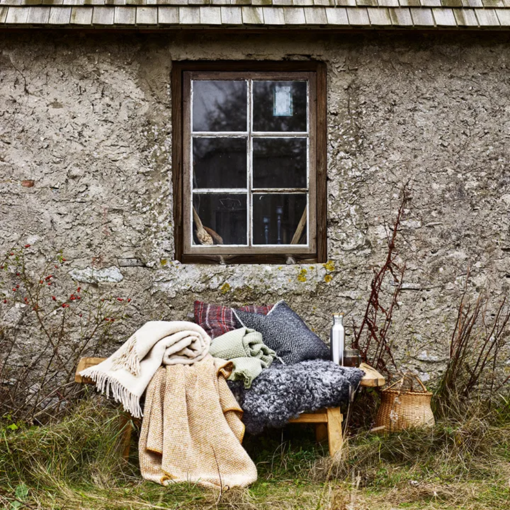 A wooden bench with blankets and cushions sits outside a weathered stone building under a window; a lantern and thermos are nearby.
