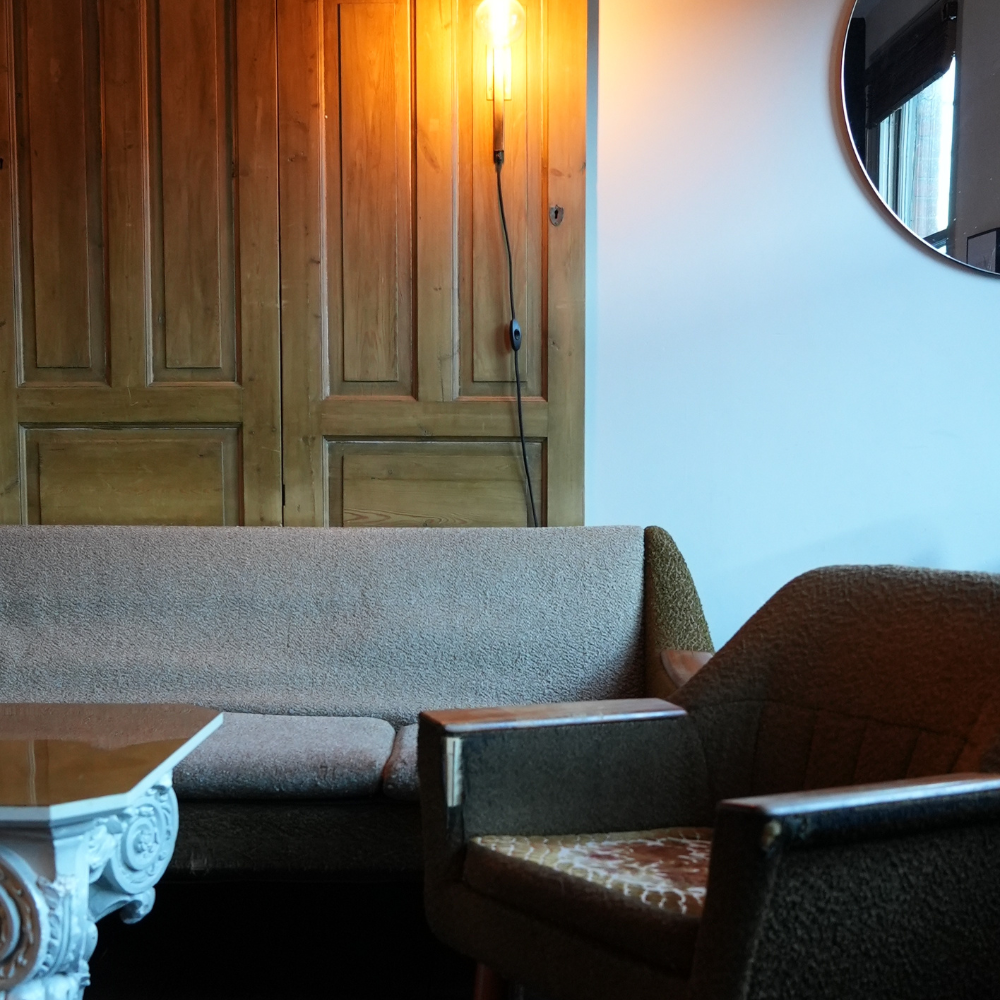 A cozy living room with a beige sofa, vintage armchair, wooden wall panels, a round mirror, a coffee table with white ornate legs, and a lit exposed bulb on the wall.