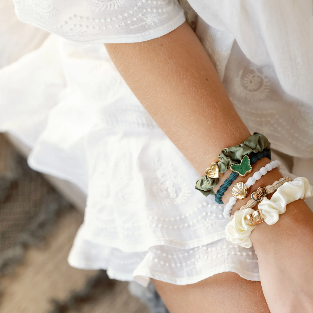 A person wearing a white embroidered dress has several decorative scrunchies and bracelets with charms and beads on their wrist. The background includes a textured rug.