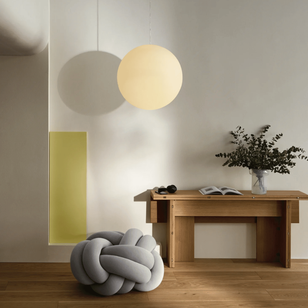 A modern, minimal room with a round hanging lamp, a wooden desk with a vase of greenery, and a gray knot cushion on a wooden floor. A yellow wall accent is visible in the background.
