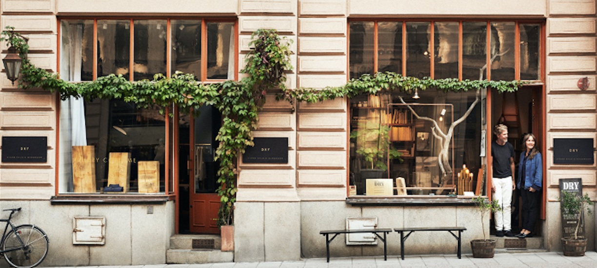 A stylish storefront with large windows, climbing greenery, a bench, and a bicycle; a man and woman stand in the doorway, looking out onto the street.