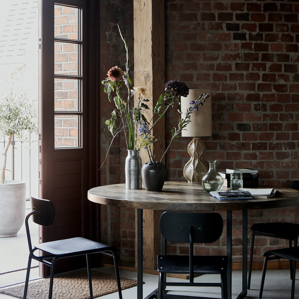 A round wooden table with black chairs sits by an open door. Vases with tall flowers and a sculptural glass piece decorate the table. The room has a brick wall and natural light.