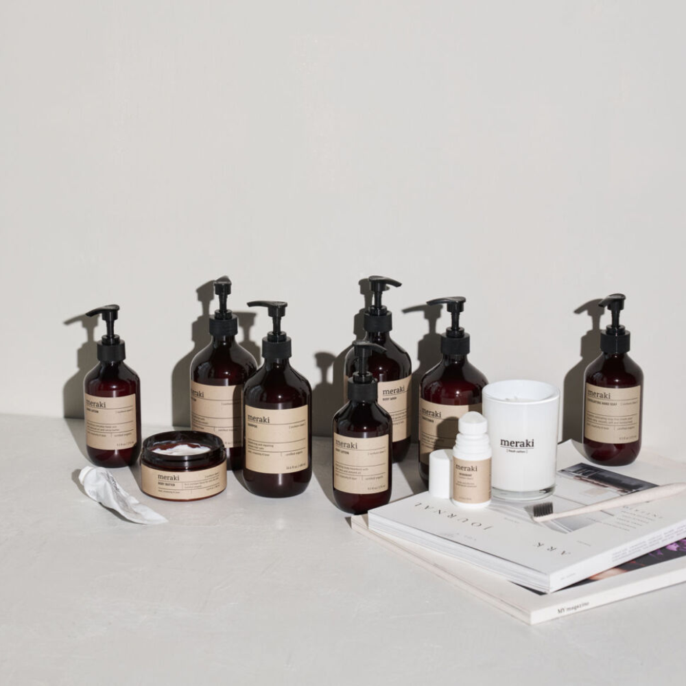 A group of amber-colored Meraki skincare bottles and jars with beige labels arranged on a white surface next to a candle and stacked magazines.