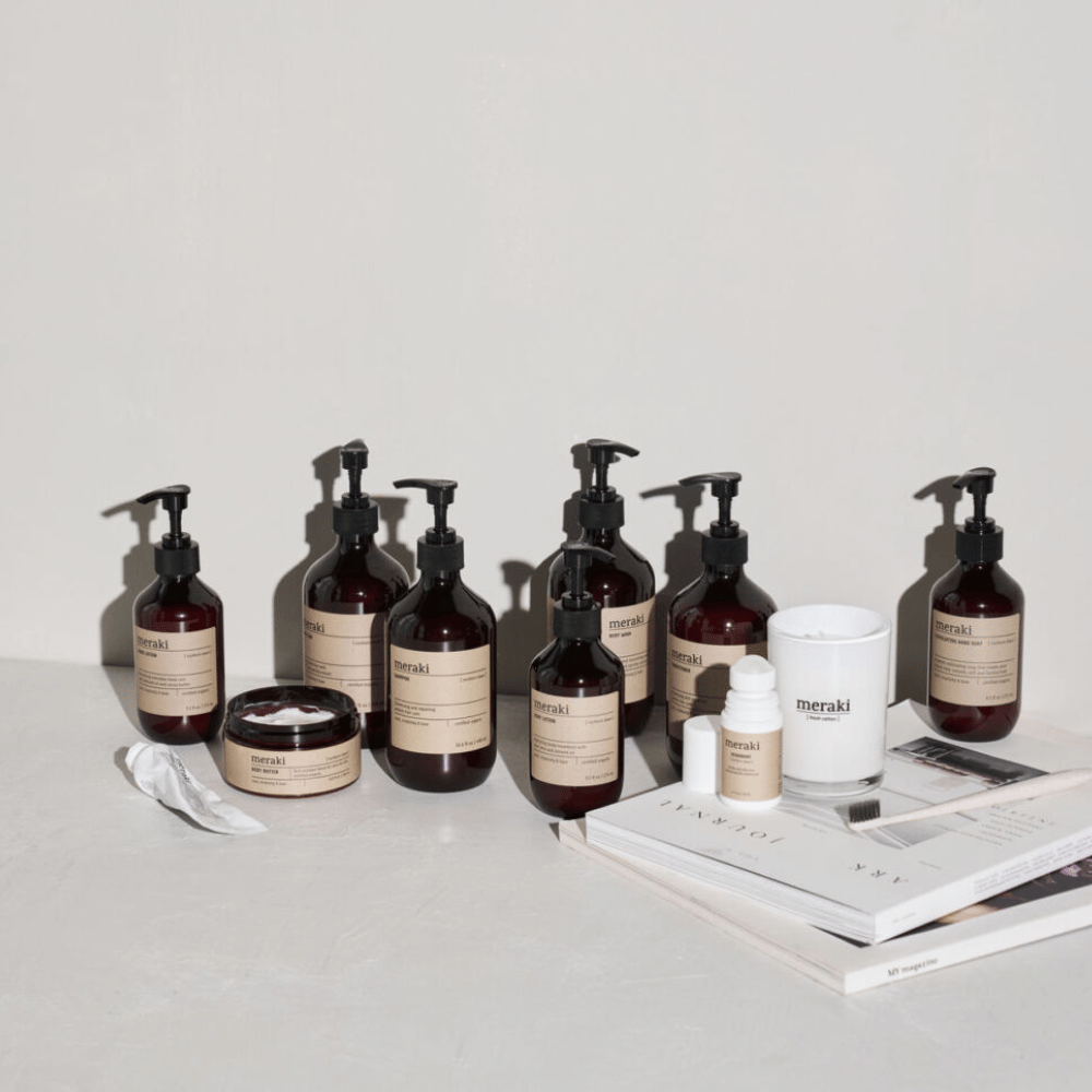A group of amber-colored Meraki skincare bottles and jars with beige labels arranged on a white surface next to a candle and stacked magazines.