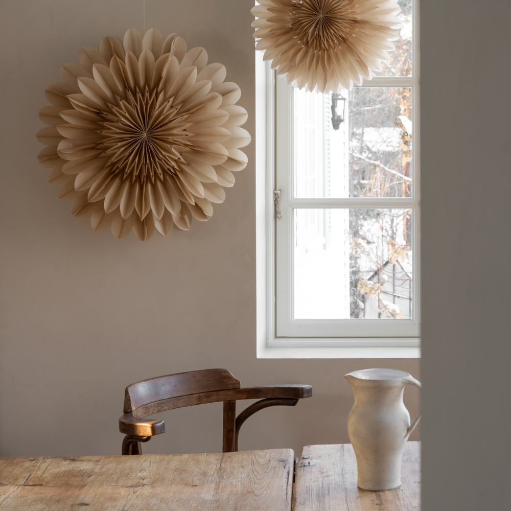 A minimalist room with a wooden table, a ceramic pitcher, a wooden chair, and two large, beige paper ornaments hanging near a bright window.