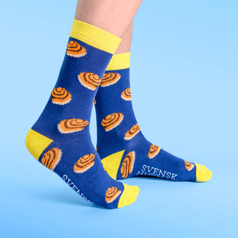 A person wearing blue socks with yellow toes, heels, and cuffs, decorated with orange cinnamon roll patterns. The word SVENSK is visible on the side. The background is light blue.