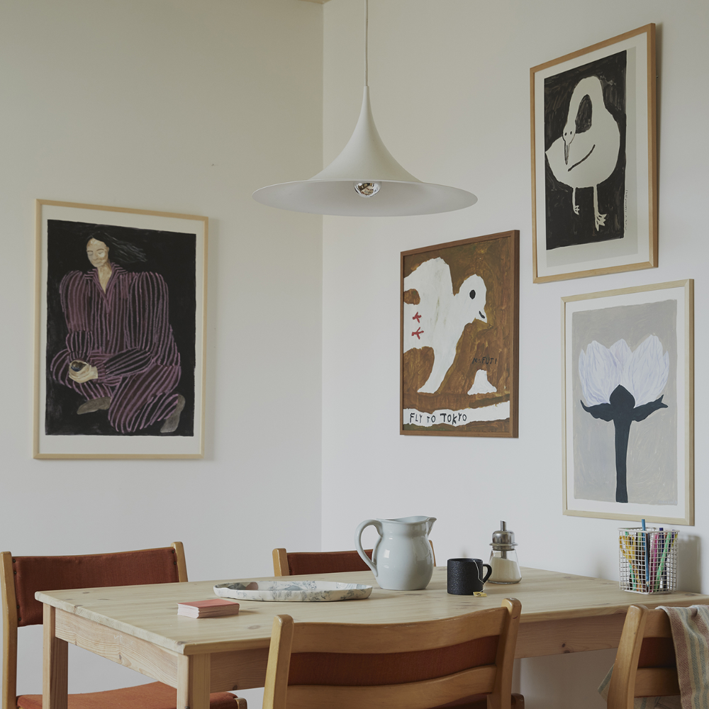 A dining area with a wooden table, red chairs, a ceramic pitcher, and four framed artworks on the wall, including abstract figures, birds, and a flower. A white pendant lamp hangs above the table.