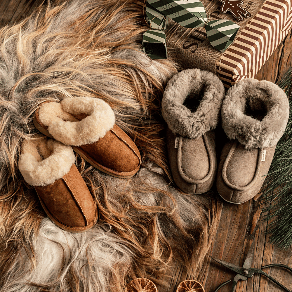 Two pairs of cozy, fur-lined slippers—one brown and one gray—rest on a plush rug beside a wrapped gift, greenery, and dried orange slices on a rustic wooden floor.