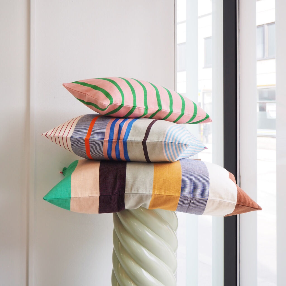 Three colorful striped pillows are stacked on top of a twisted, light green pedestal near a window, with sunlight streaming into the modern, minimalistic room.