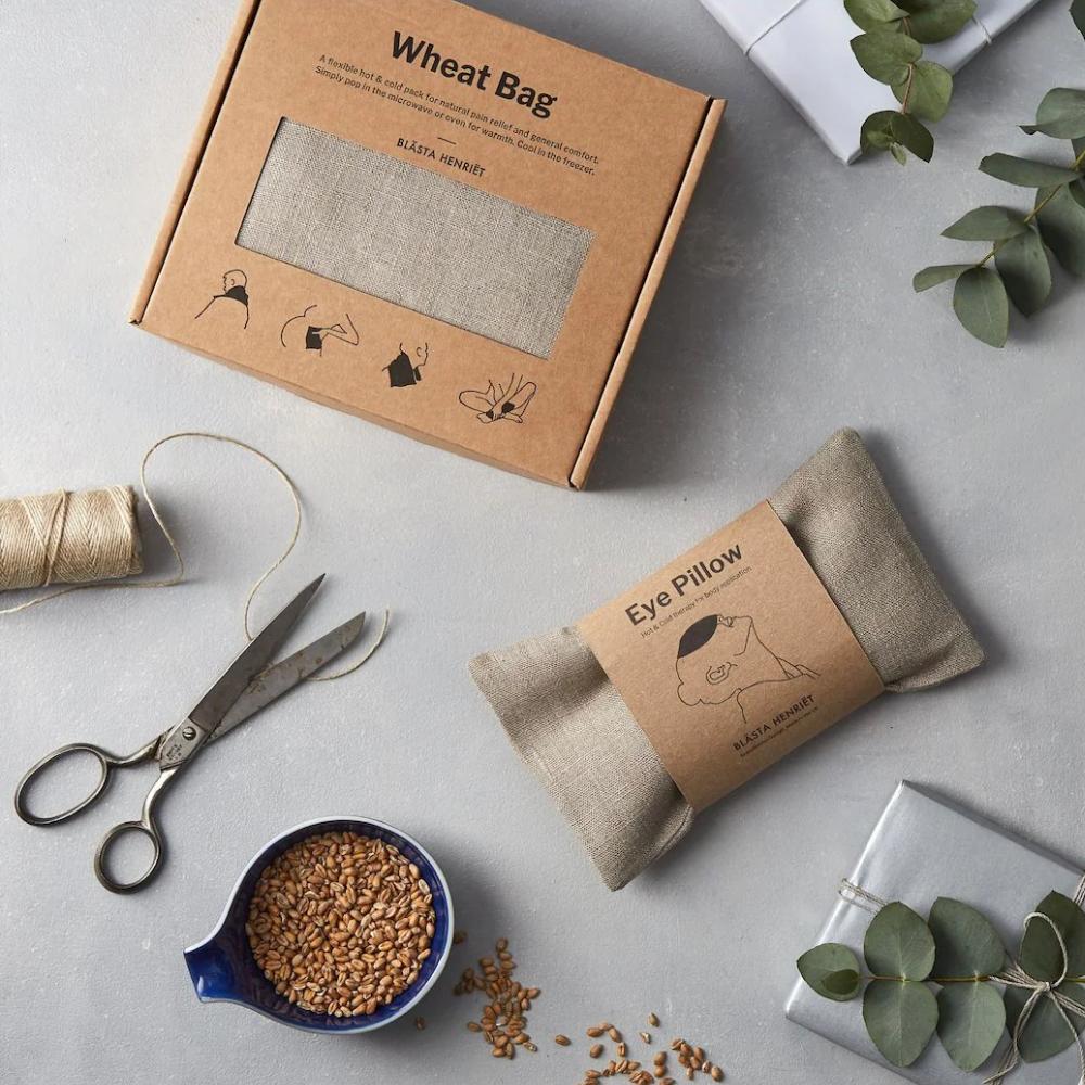 A boxed wheat bag, a matching eye pillow, loose wheat grains, scissors, twine, eucalyptus sprigs, and wrapped gifts arranged on a light gray surface.