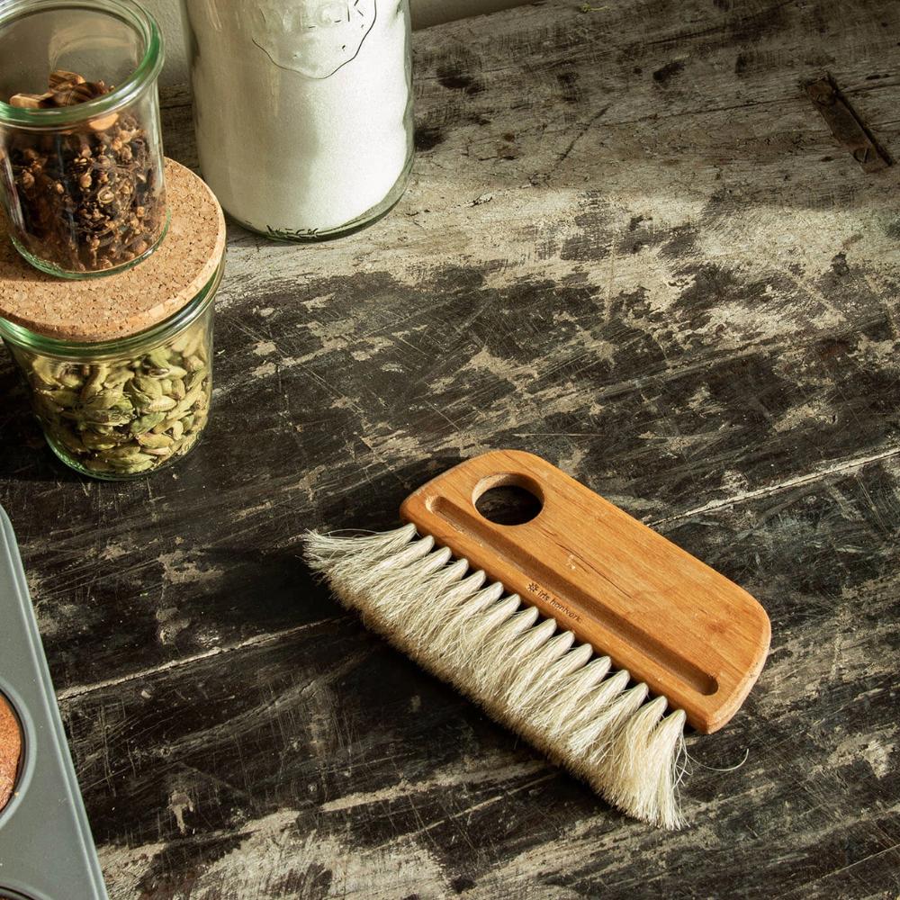 A small wooden brush with white bristles lies on a rustic, worn wooden surface. Nearby are glass jars with cork lids containing spices and a larger jar filled with white powder, possibly sugar.