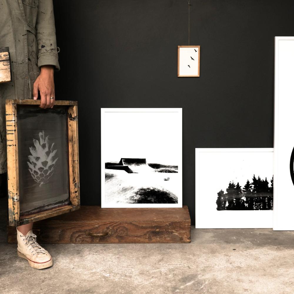 A person in sneakers and a long coat holds a wooden frame with a leaf print, standing beside minimalist black-and-white art prints against a dark wall.