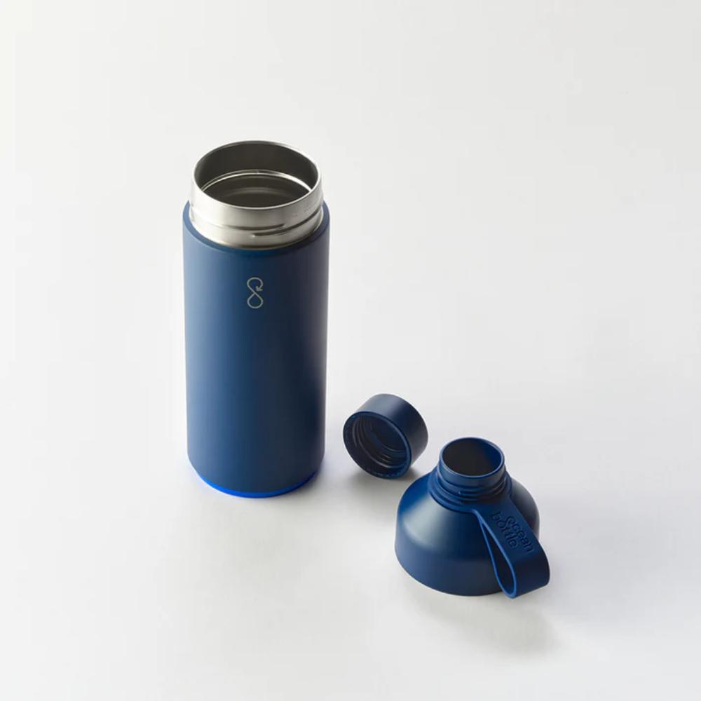 A blue reusable water bottle with a removable cap and a wide mouth lid, both placed on a white surface next to the bottle. The bottle has a silver interior and a minimalist design.