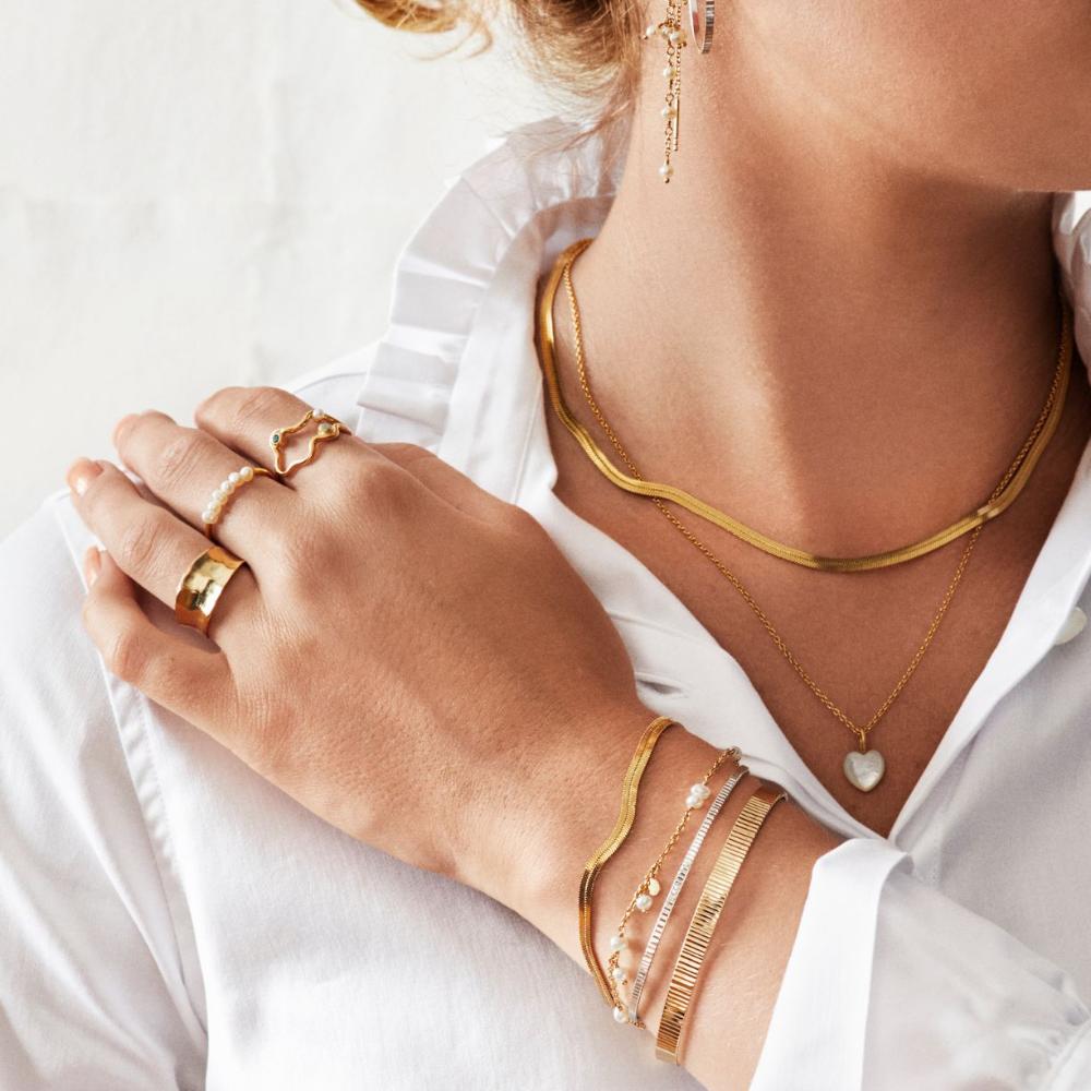 A woman wearing layered gold necklaces, bracelets, and rings over a white blouse. The jewelry includes a heart pendant and pearl-accented pieces. Only her hand, neck, and part of her face are visible.