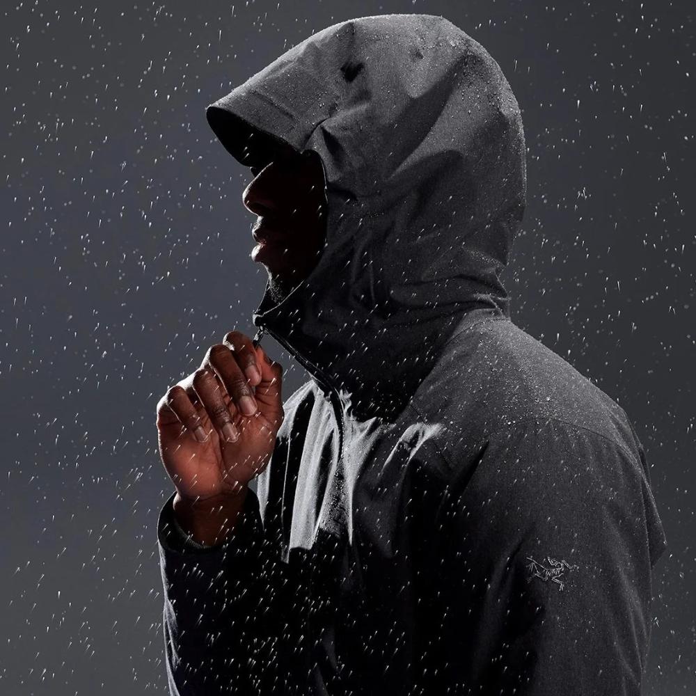 A person wearing a hooded rain jacket stands in the rain, shown from the side with droplets visible on the jacket.