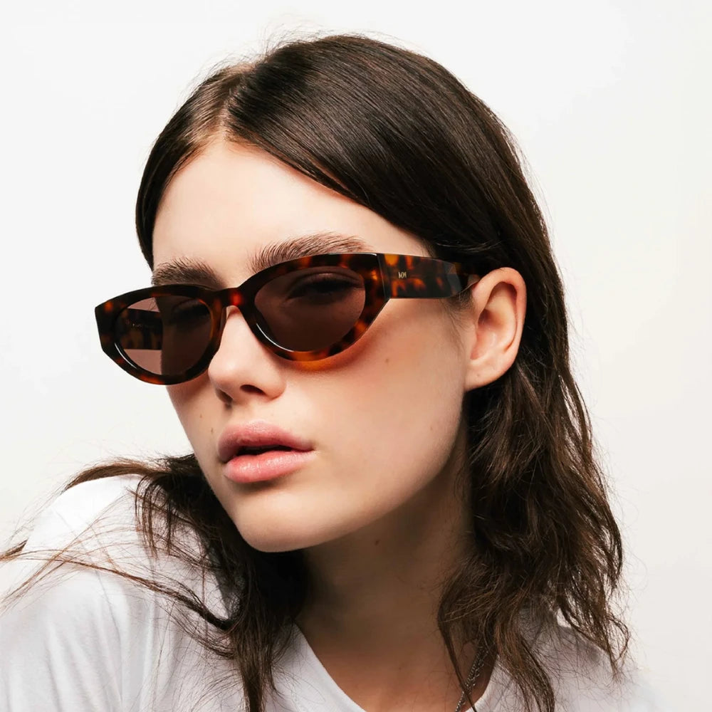 A person with long, dark hair wearing tortoiseshell cat-eye sunglasses and a white shirt poses against a plain white background, looking slightly to the side with a neutral expression.