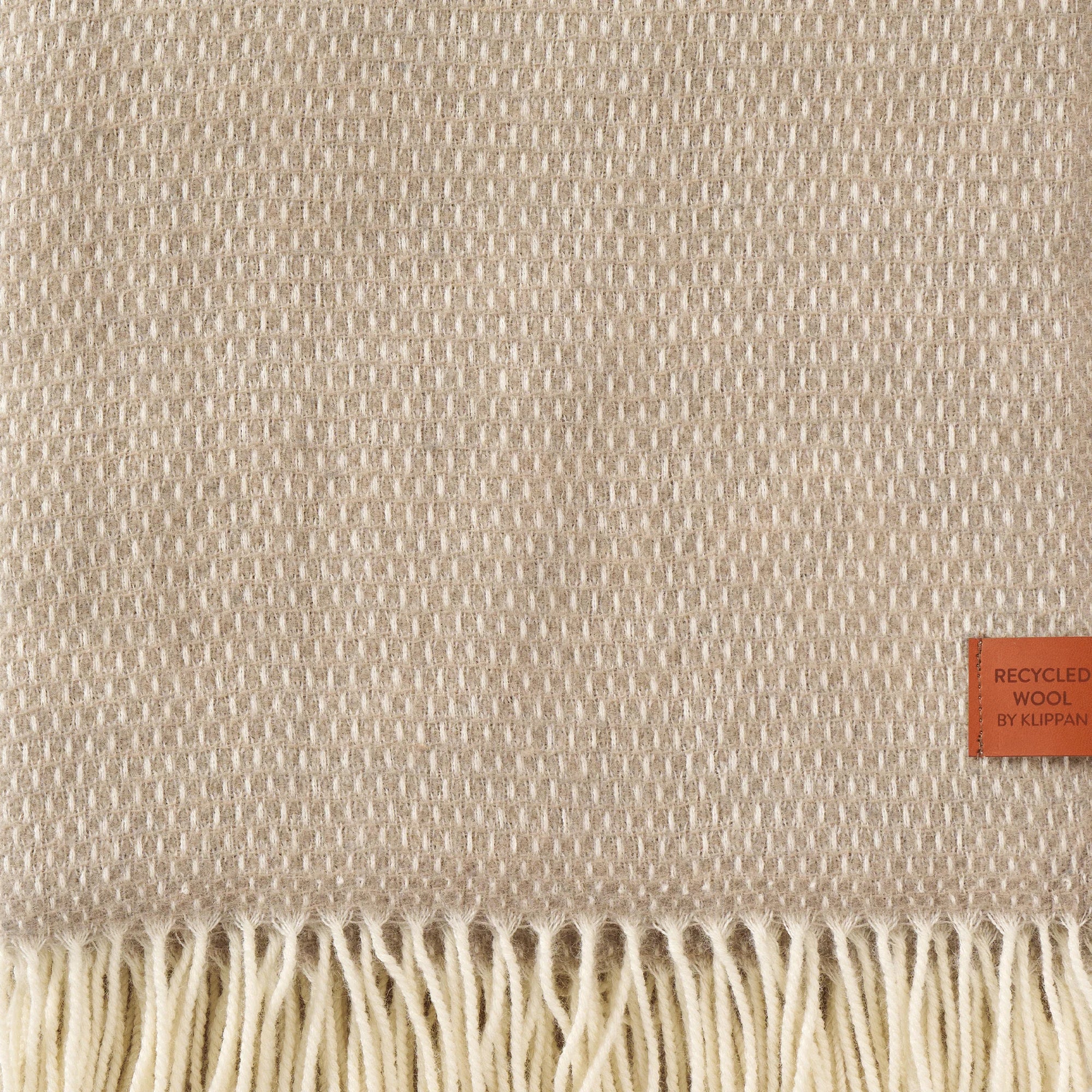 The Hugo Recycled Merino & New Merino Brushed Throw in Taupe by Klippan features a beige textured finish, a bottom fringe, and a Recycled Wool tag, showcasing the quality of recycled wool throws.