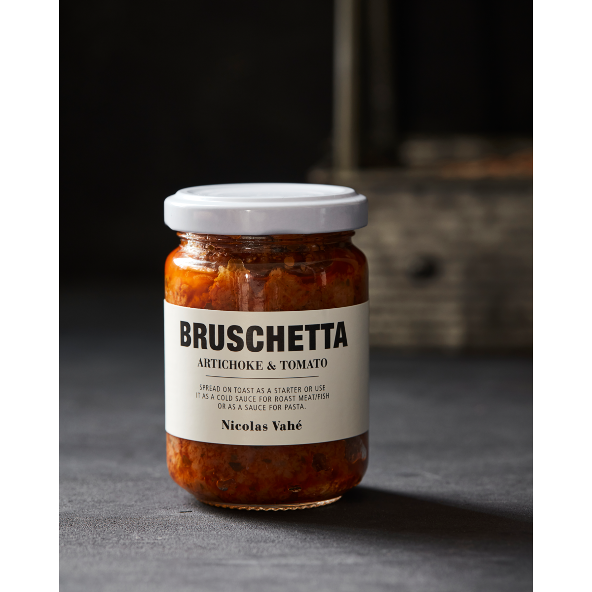 A jar of Nicolas Vahé Bruschetta, Artichoke & Tomato—ideal as a pasta sauce—rests on a dark surface against a blurred background.