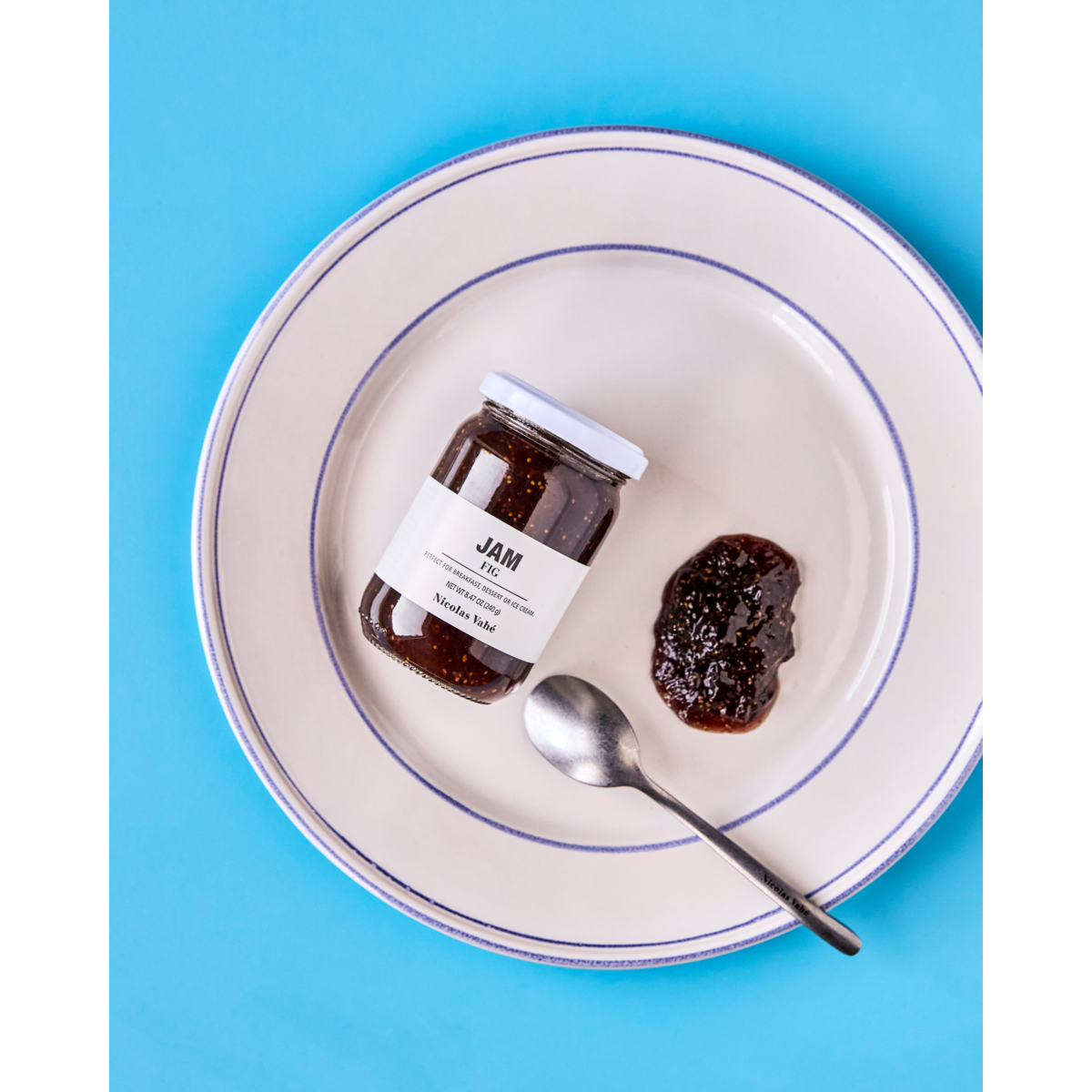 A jar of Nicolas Vahé Jam, Fig, a spoonful of jam, and a spoon rest on a white plate with a blue rim against a blue background—ideal for your cheese table.