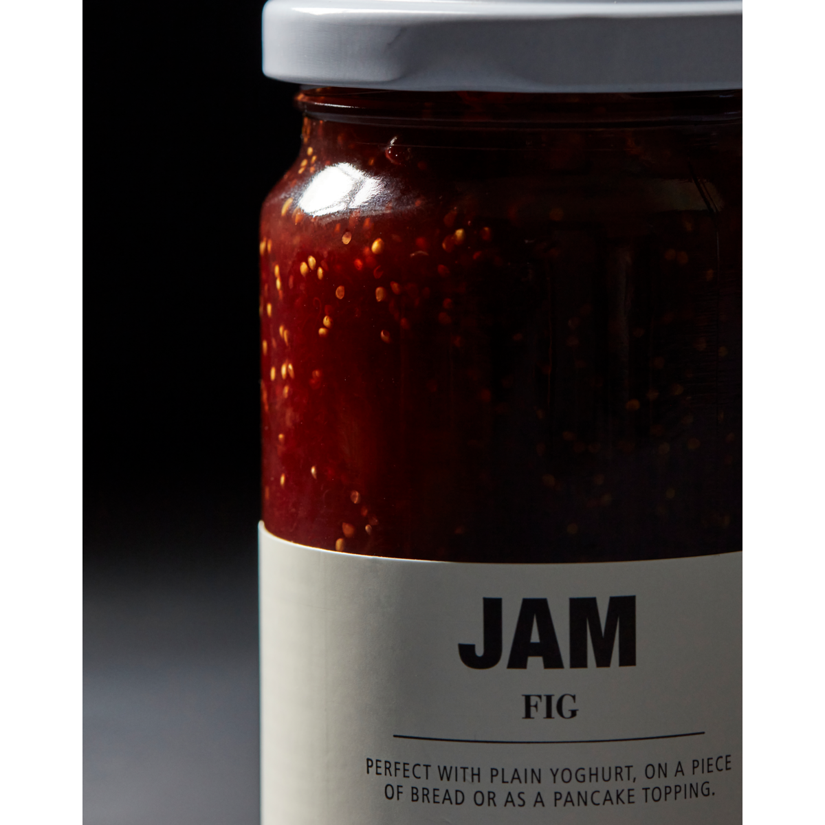 A jar of Nicolas Vahé Jam, Fig with a white lid sits against a dark background. The label recommends serving it with yogurt, bread, pancakes, or adding it to your next cheese board.