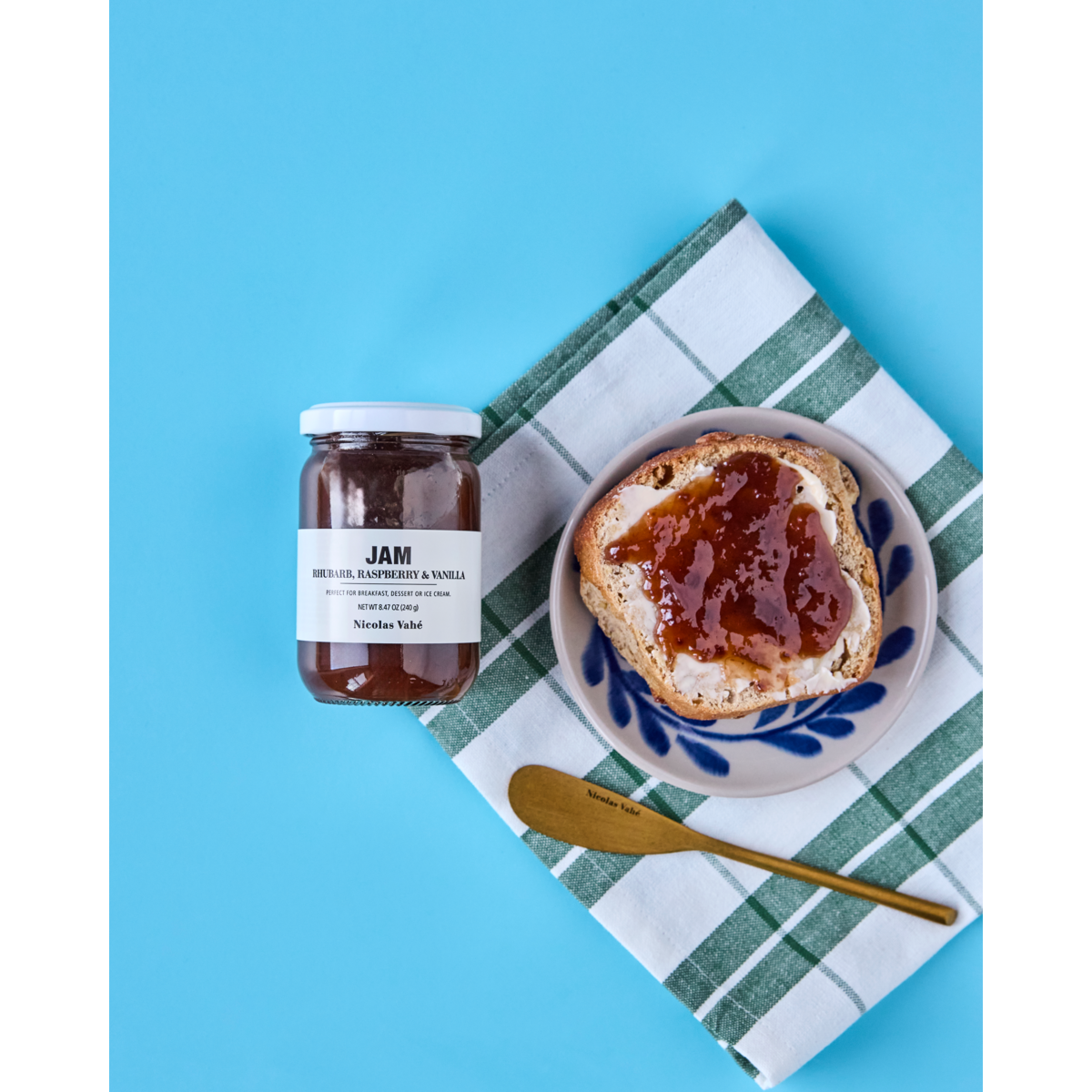 A jar of Nicolas Vahé Jam, Rhubarb, Raspberry & Vanilla and a slice of toast with jam on a plate beside a gold knife, all set on a green and white checkered cloth against a blue background.