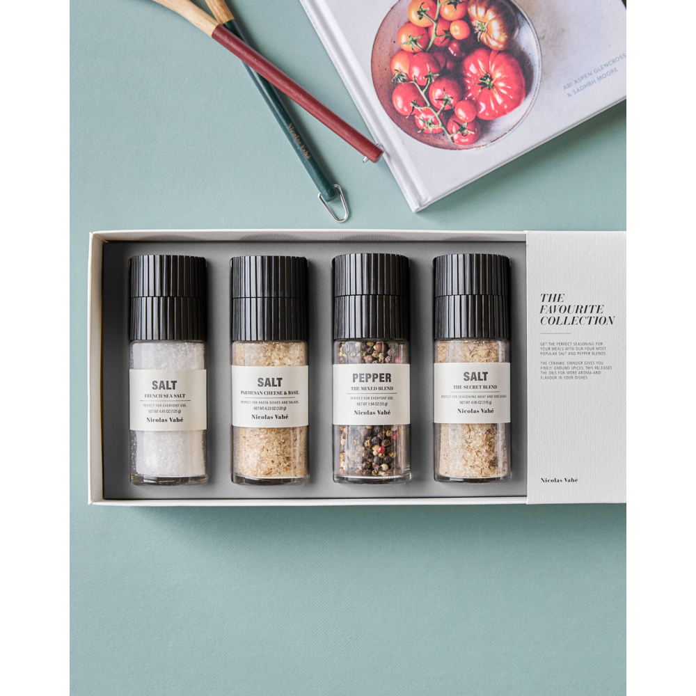 A box set from Nicolas Vahé—called the Gift Box, Favourite Collection, featuring French Sea Salt, Parmesan Cheese & Basil Salt, The Secret Blend Salt, and The Mixed Pepper Blend—rests on a light blue surface, with a cookbook and utensils partially visible in the background. Each grinder includes a premium ceramic mechanism for optimal freshness.