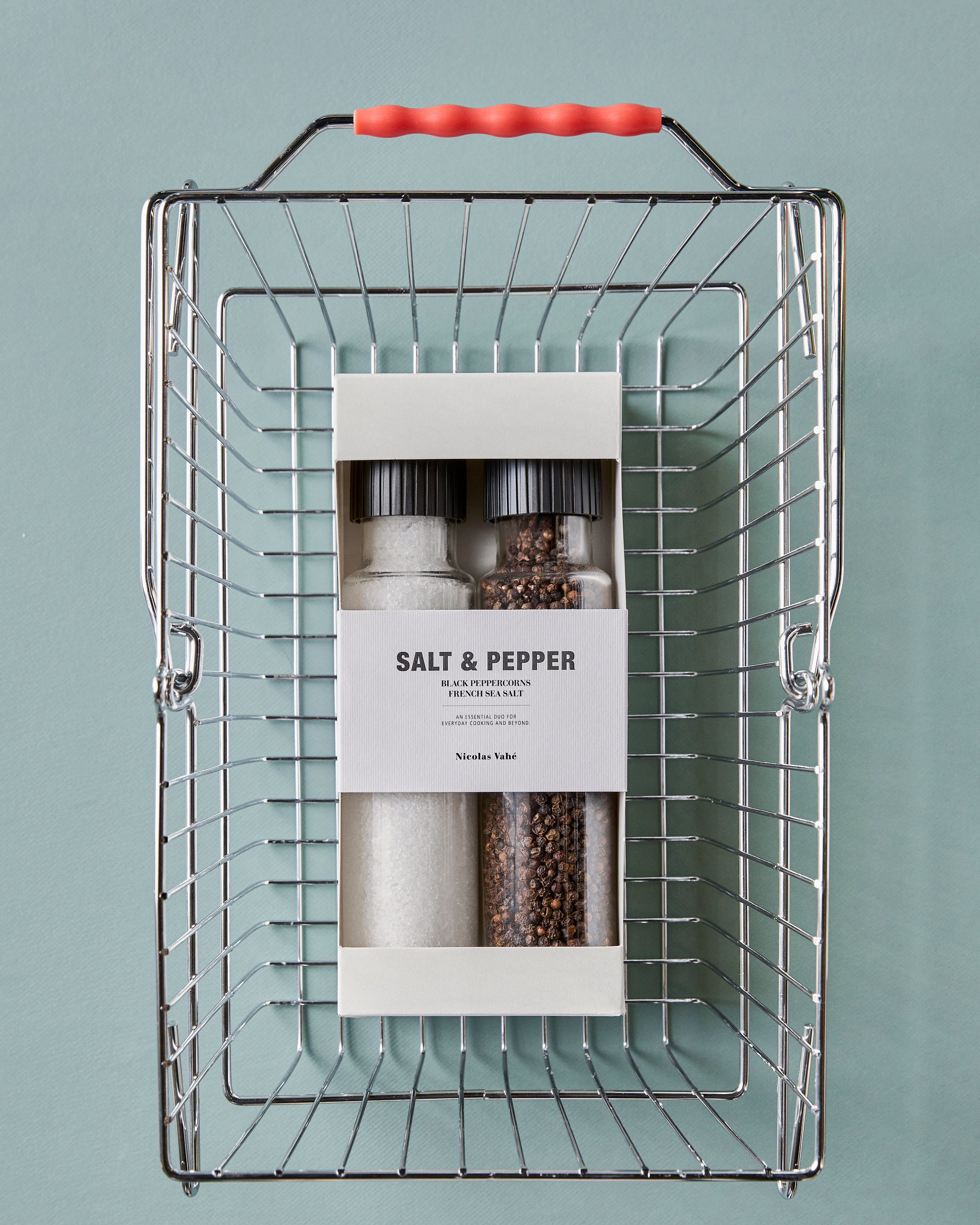 A metal shopping basket holds a Nicolas Vahé Gift Box, Sea Salt & Black Pepper, Tall, with salt and pepper grinders in clear bottles featuring black caps and white labels, set against a light blue background.