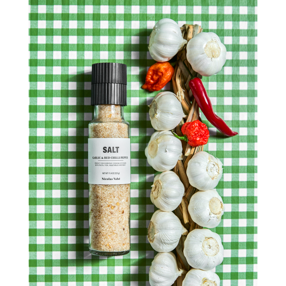 A bottle of Nicolas Vahé Salt, Garlic & Red Chilli Pepper sits beside a braid of garlic bulbs and some red chili peppers on a green and white checkered tablecloth.