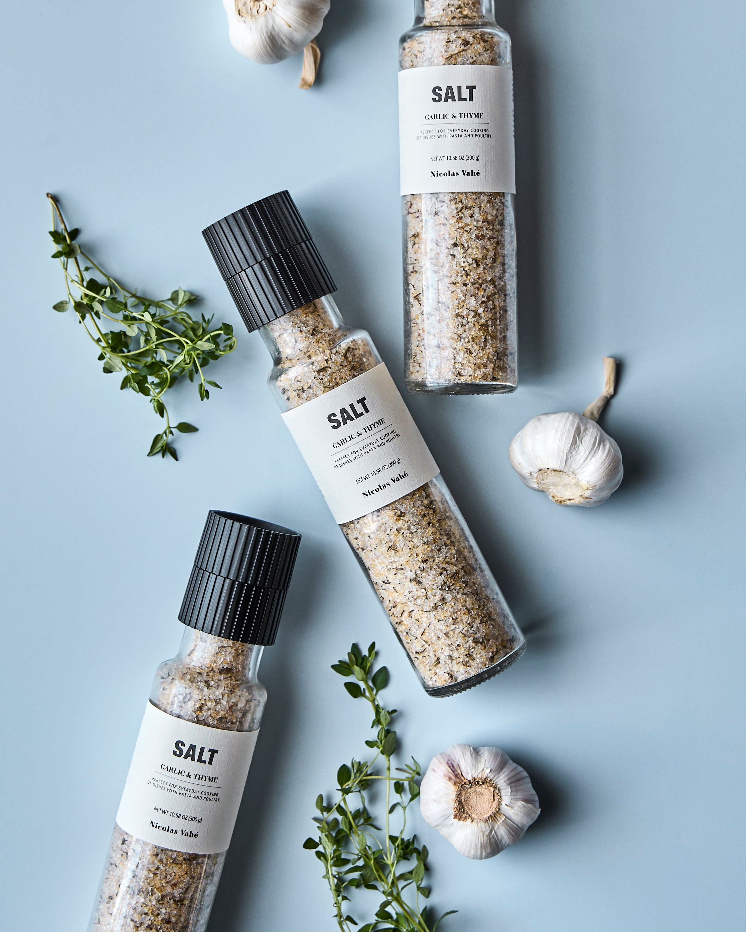 Three Nicolas Vahé "Salt, Garlic & Thyme" glass grinders with black caps are arranged on a light blue surface, surrounded by sprigs of thyme and whole garlic bulbs. The labels highlight their artisanal blend of premium salt infused with garlic and thyme.