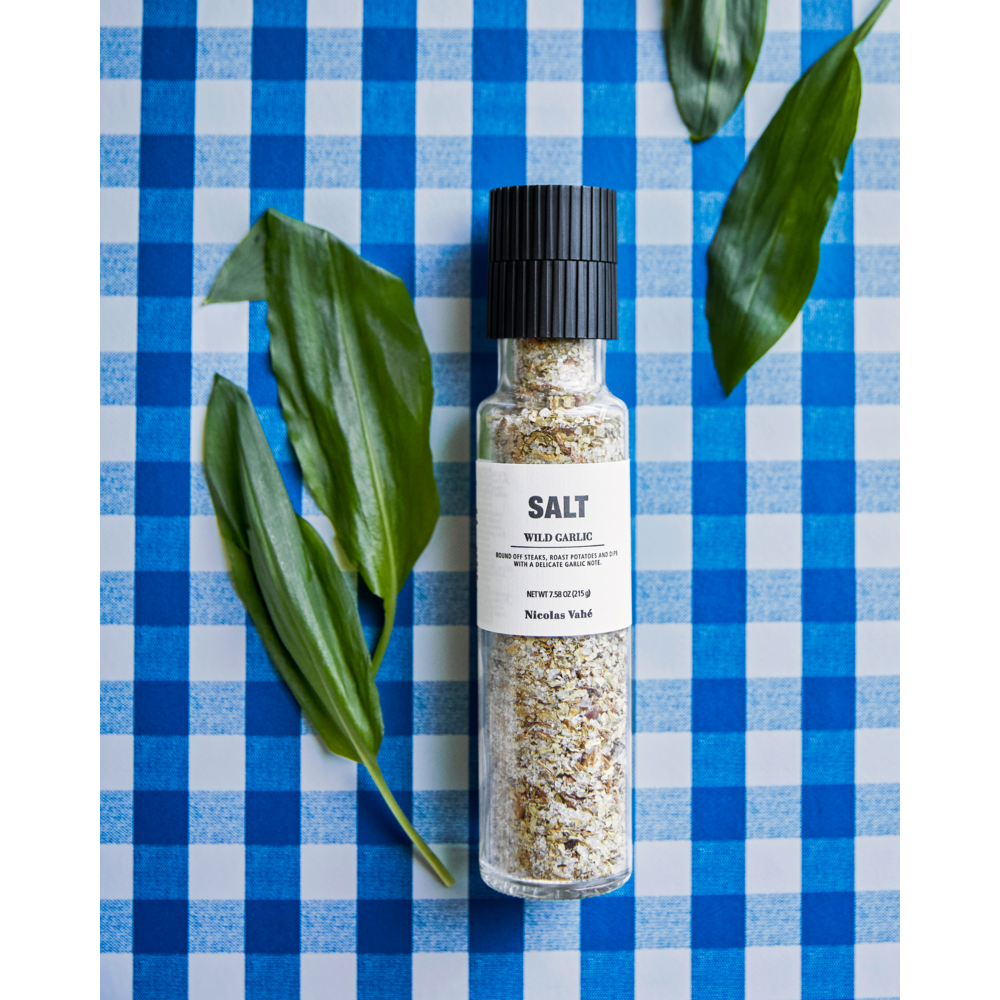 A bottle of Nicolas Vahé Salt, Wild Garlic with a ceramic grinder rests on a blue and white checkered tablecloth, accompanied by three green leaves placed beside it.