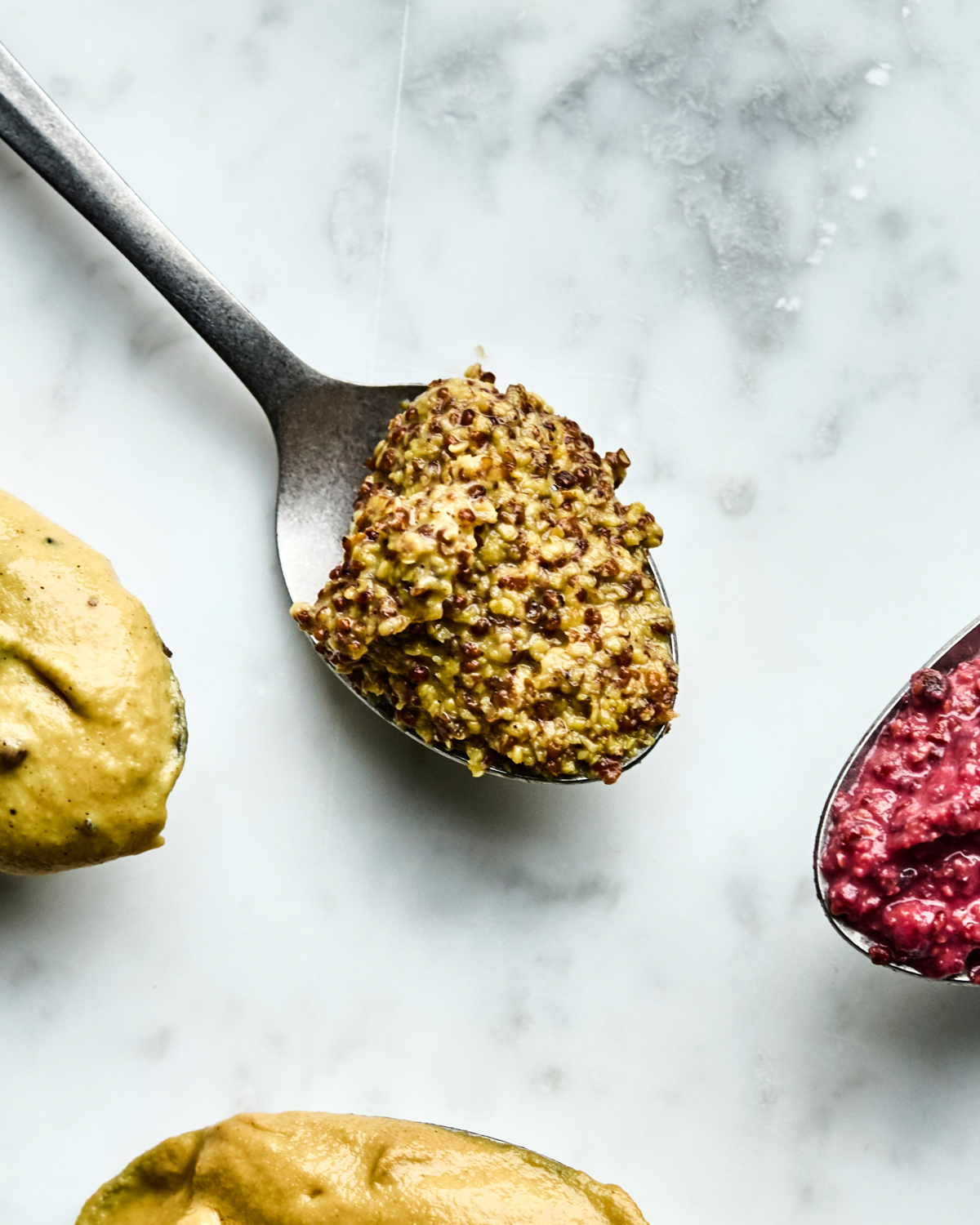 A close-up of three Nicolas Vahé mustards—Mustard, Honey & Whole Grain—on a marble surface, each spoon displaying a different variety.