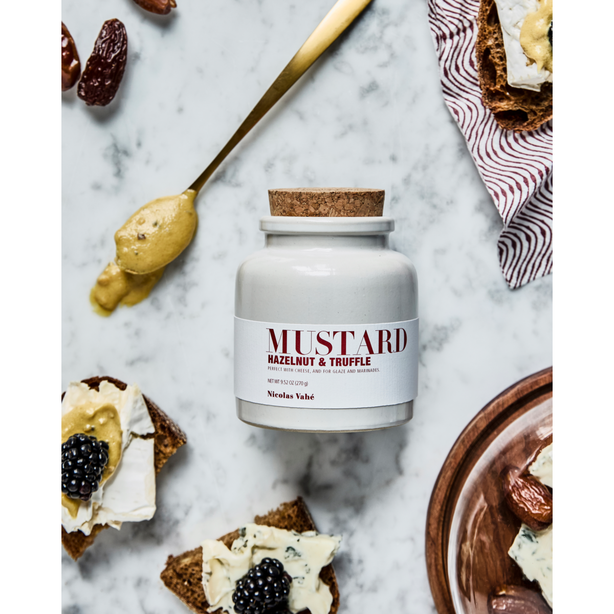 A jar of Nicolas Vahé Mustard, Hazelnut & Truffle sits on marble, with a spoonful of the gourmet mustard and cheese-topped bread with blackberries nearby—a festive condiment perfect for special occasions.