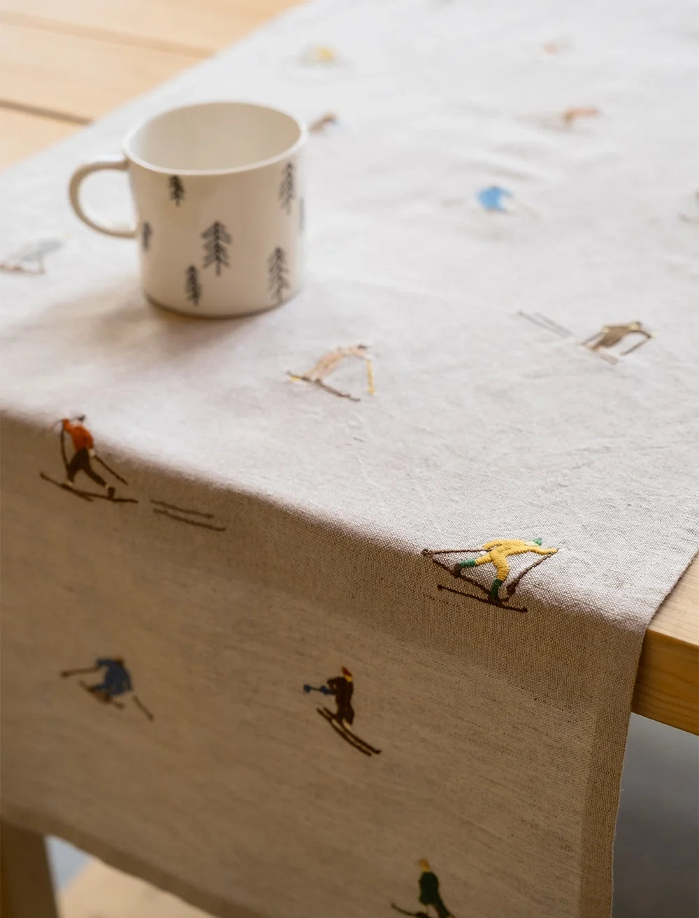 A white mug with tree illustrations rests on the Fine Little Day Skiers Embroidered Table Runner, a beige linen cotton textile adorned with colorful skiers in various poses, adding a charming touch to winter table decor.