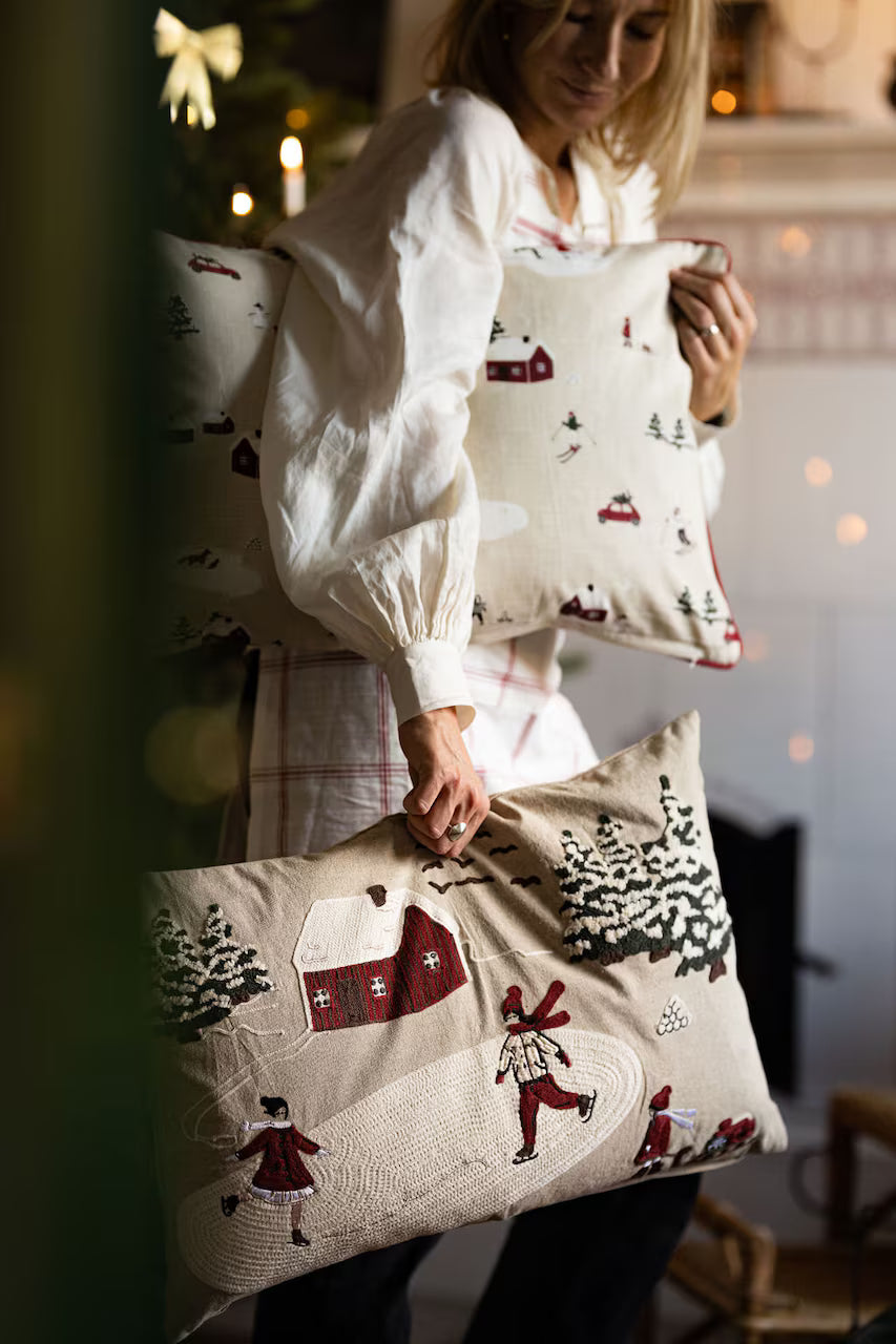 A person holds two Strömshaga Cushion Cover Vinterland Embroidered (40 x 60 cm) with winter village scenes—skaters and snowy houses—in a cozy room, creating the perfect winter wonderland cushion display.