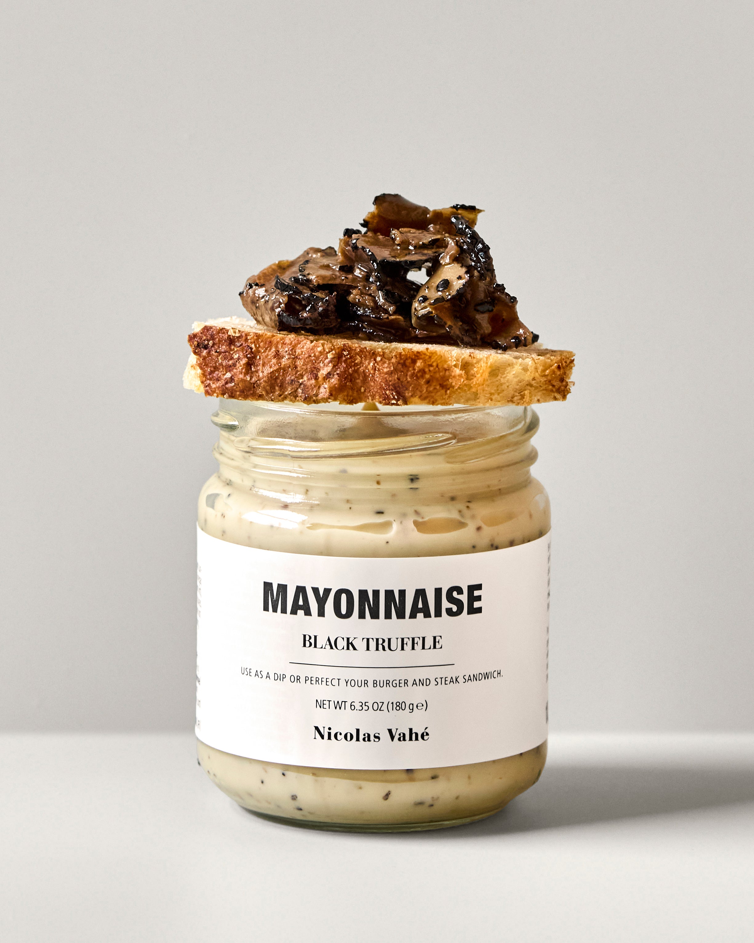A jar of Nicolas Vahé Mayonnaise, Black Truffle stands against a plain light background, its lid balancing a slice of bread topped with sautéed mushrooms.
