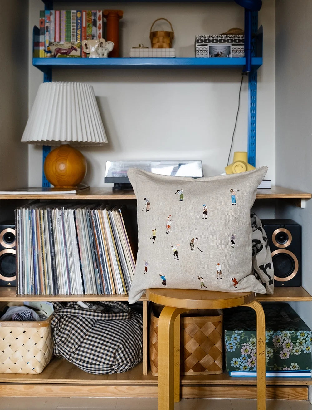 A cozy shelf setup with records, speakers, baskets, books, a lamp, and the Fine Little Day Golfers Embroidered Cushion Cover in unbleached linen with small colorful figures—all arranged neatly on and under wooden shelves.