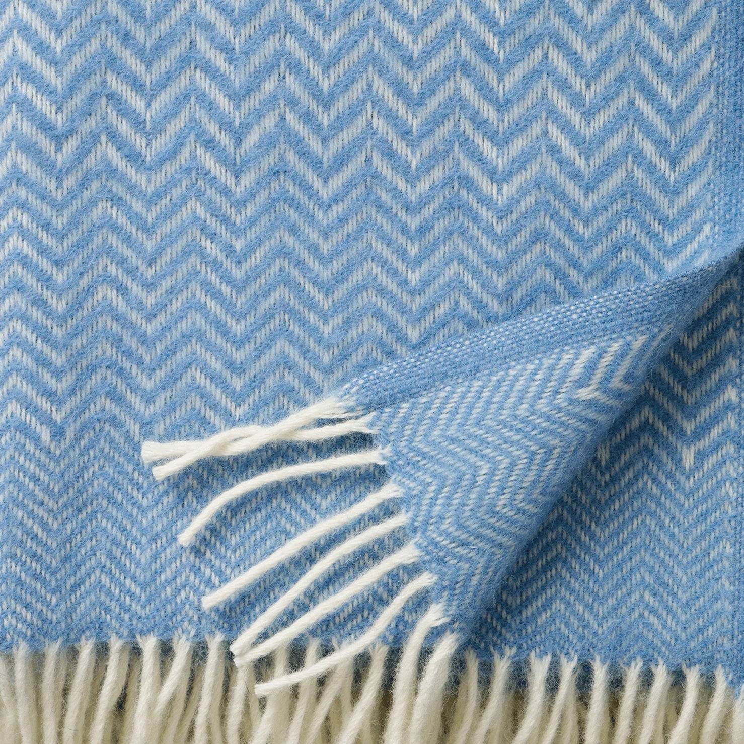 The Zigzag Throw in Lambswool, Infinity Blue by Klippan features a stylish blue and white herringbone pattern with fringed edges—perfect for adding a cozy touch to your home decor.