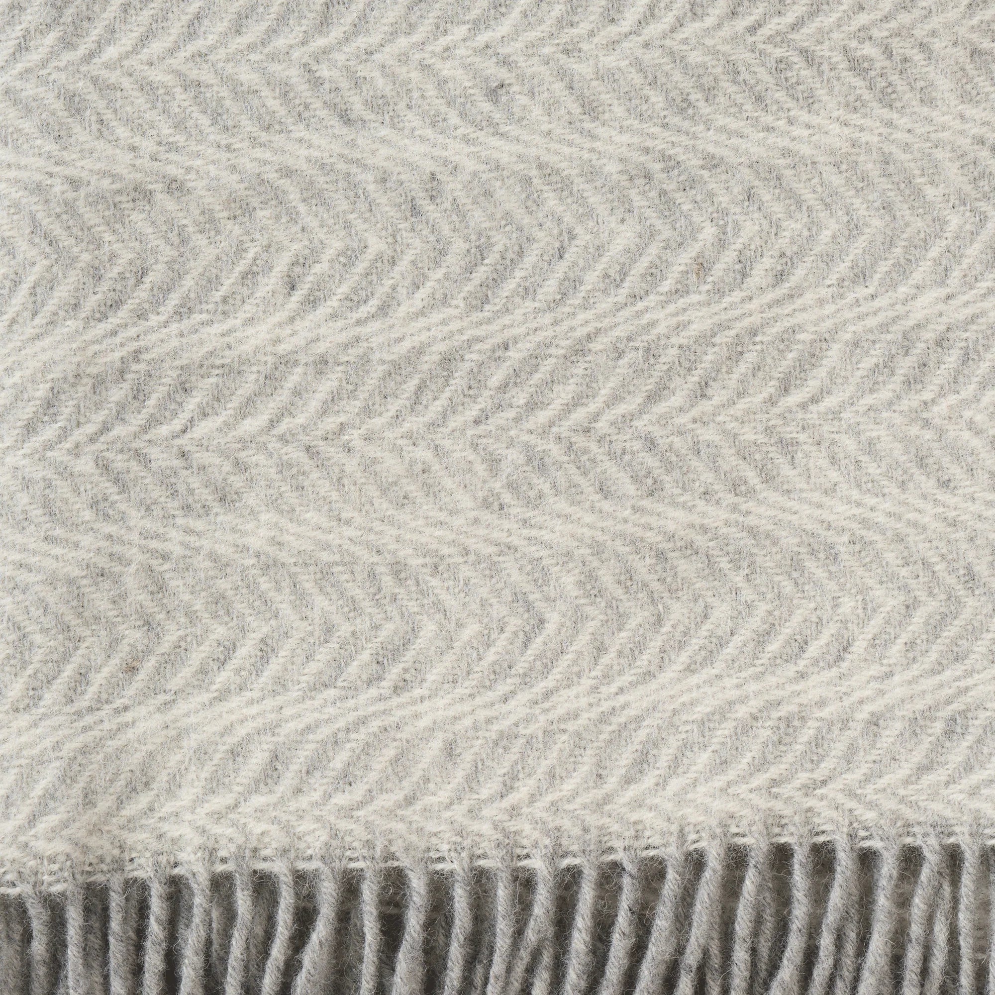 Close-up of the Klippan Alm Throw in Eco Lambswool, Ash: a light gray throw with a subtle chevron pattern and fringe along the bottom edge—an elegant addition to your home decor.