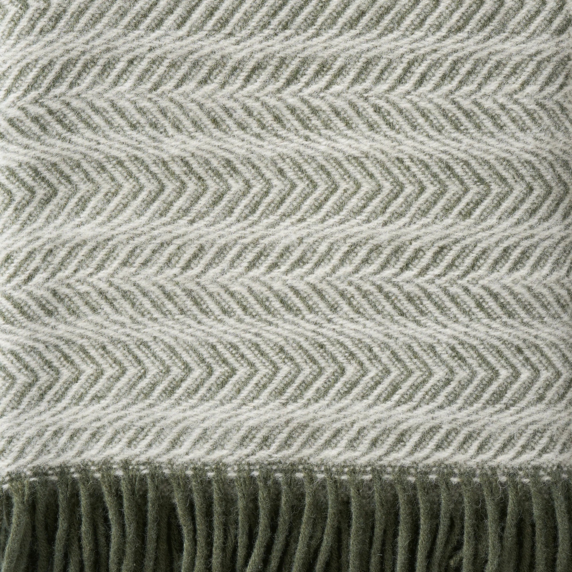 Close-up of the Klippan Alm Throw in Eco Lambswool, Pine Green, featuring a herringbone pattern in light gray and green with fringe along the edge—an elegant accent for your home.