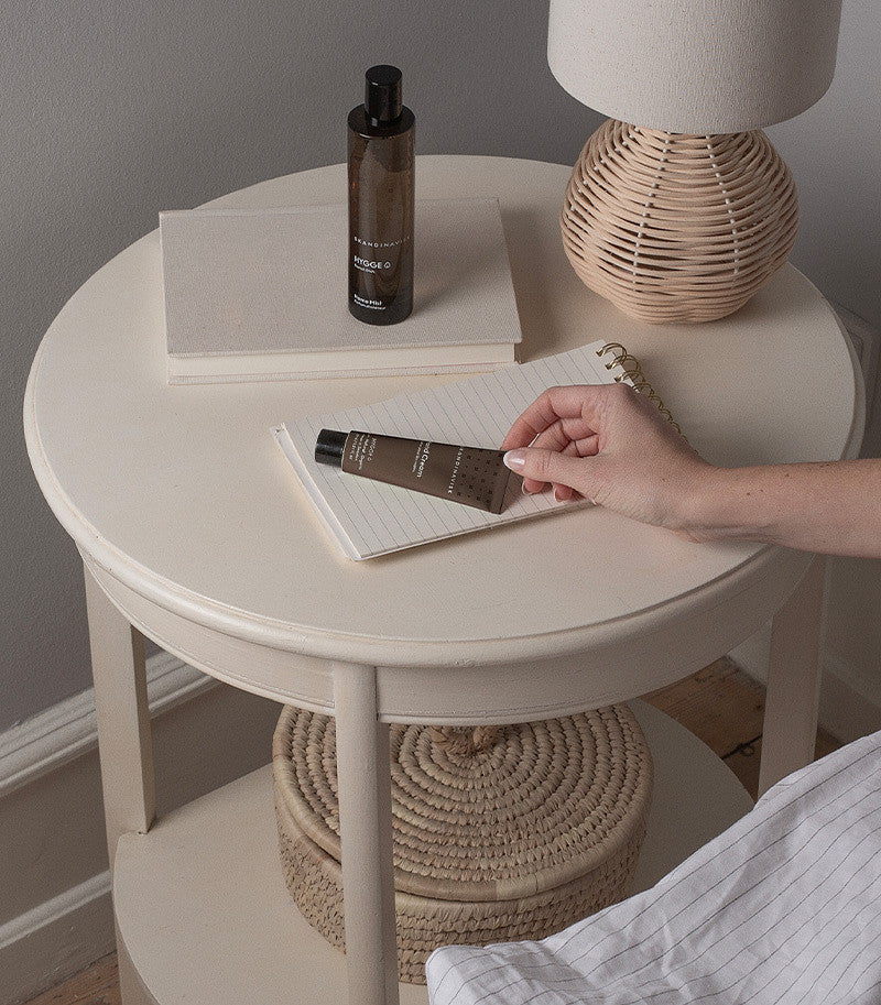 A hand reaches for a brown tube of Skandinavisk Mini Hand Cream Hygge, 30 ml, resting on a notepad atop a round beige nightstand that also displays a lamp, a closed book, and a bottle. A woven basket sits on the lower shelf, enhancing the minimalist Scandinavian skincare aesthetic.