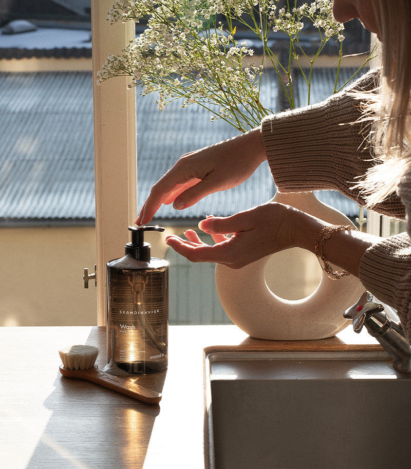 By the sink, sunlight streams through a window as someone presses the Skandinavisk Hand Wash Hygge, 500 ml pump bottle. Echoing Scandinavian hygge, a vase of white flowers and a small brush rest nearby on the counter, creating a tranquil scene while organic aloe vera in the hand wash nourishes the skin.