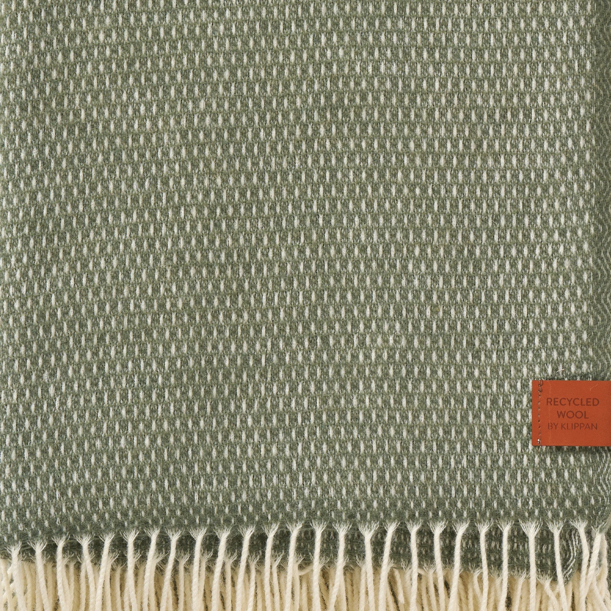 A close-up of the Klippan Hugo Recycled Merino & New Merino Brushed Throw in Dusty Green, featuring a textured weave, cream fringes, and a brown Recycled Wool by Klippan tag in the lower right corner.