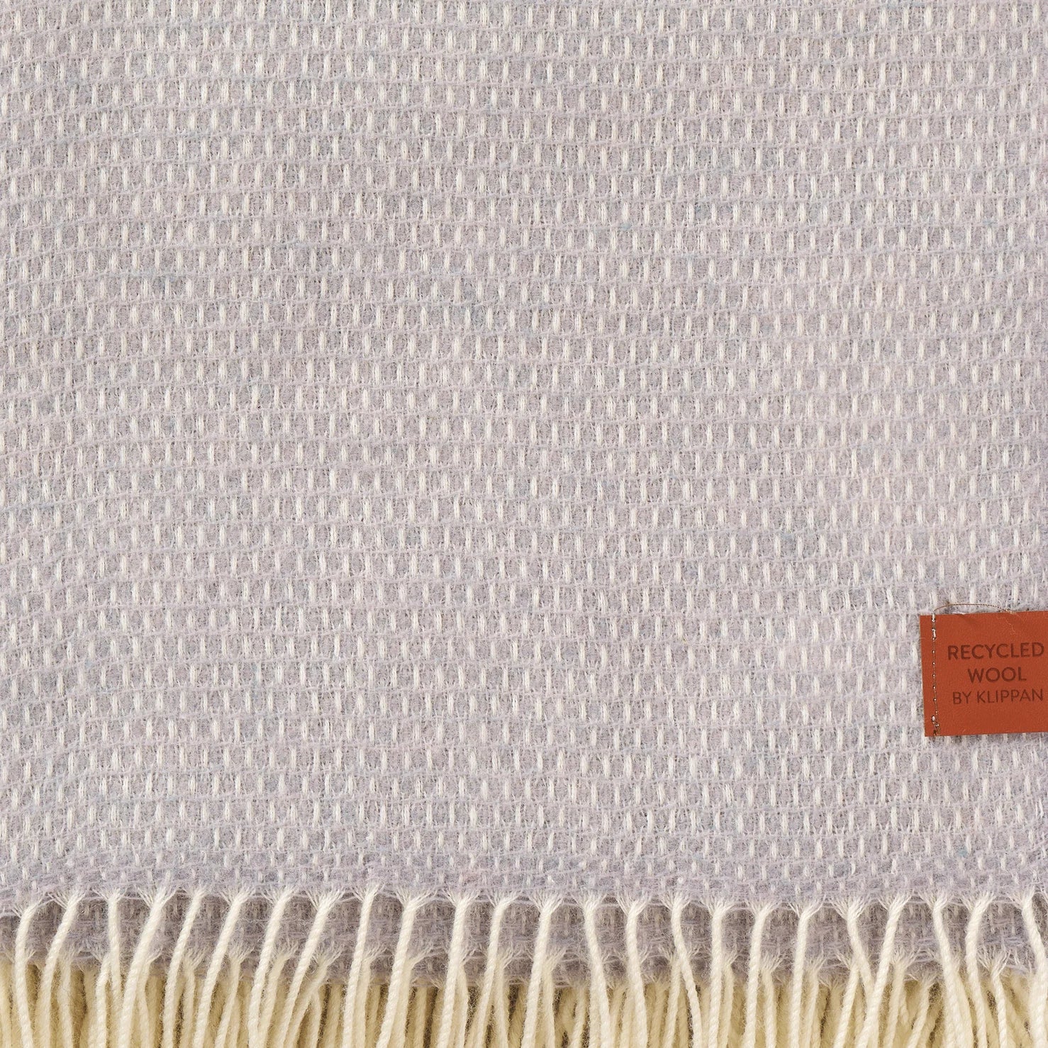 A close-up of a lavender Hugo Recycled Merino & New Merino Brushed Throw by Klippan, featuring cream fringes and a brown label that reads, Recycled Wool by Klippan.