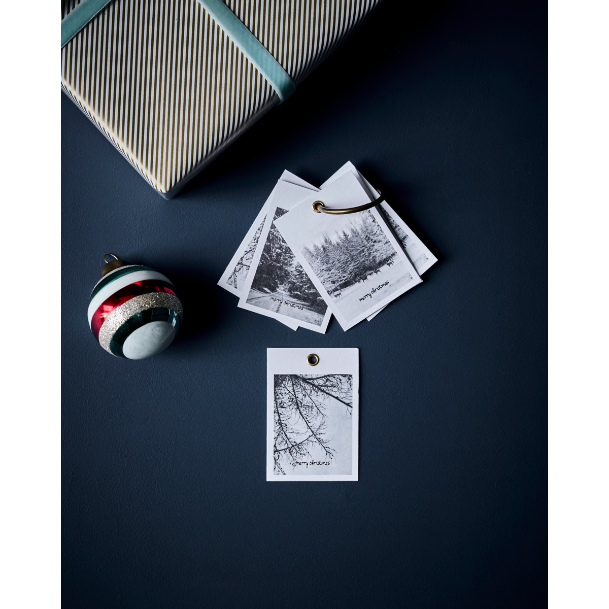 A wrapped gift with House Doctor’s Winter Gift Tags (set of 12), a striped ornament, and black-and-white winter photo cards is displayed on a dark surface.