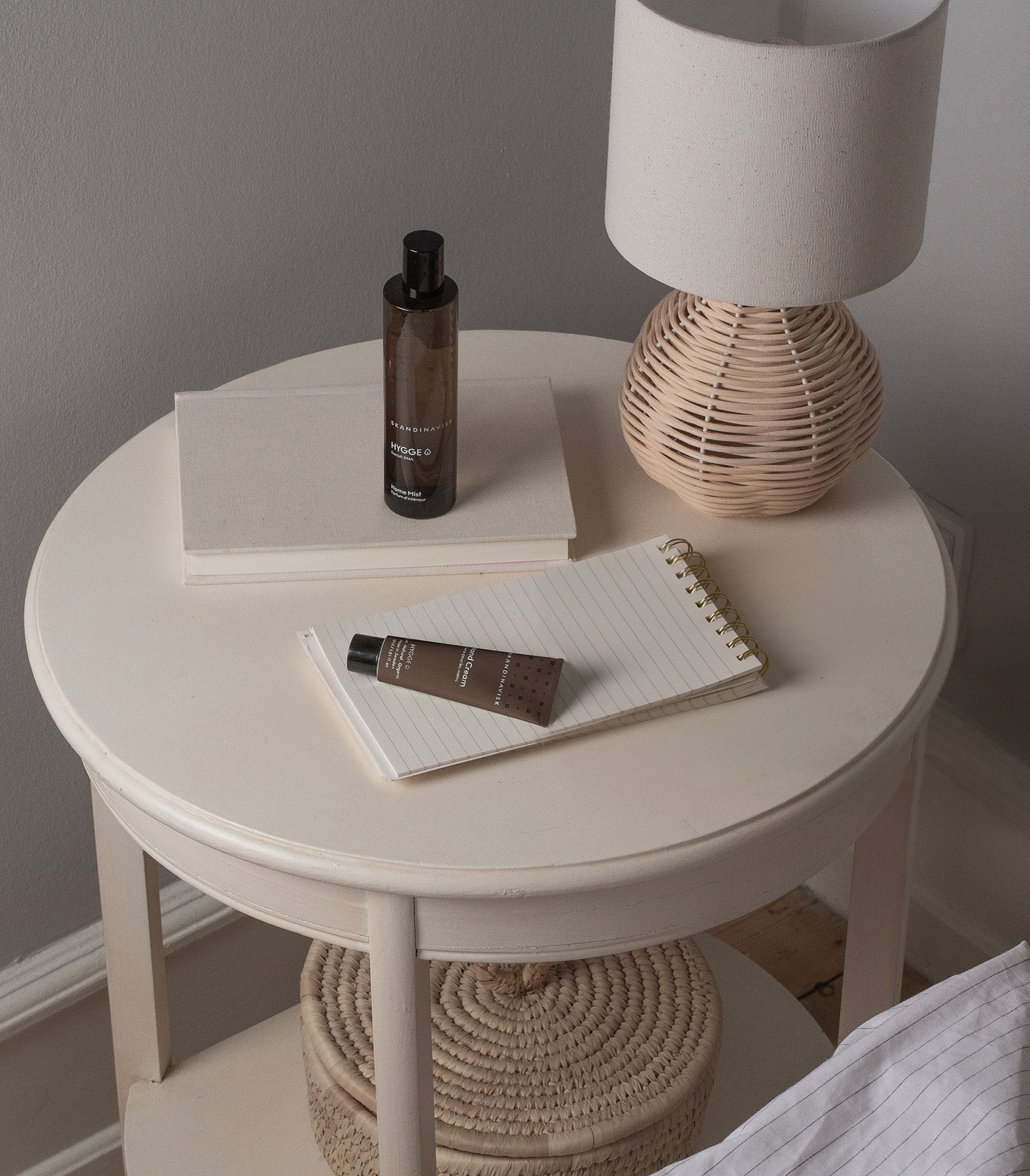 A beige round nightstand is styled with a rattan lamp, a notebook, a closed book, and Skandinavisk’s Home Mist Hygge (100 ml), arranged alongside a brown tube. Positioned beside a bed draped in a white blanket, the setting evokes Scandinavian hygge and understated elegance.