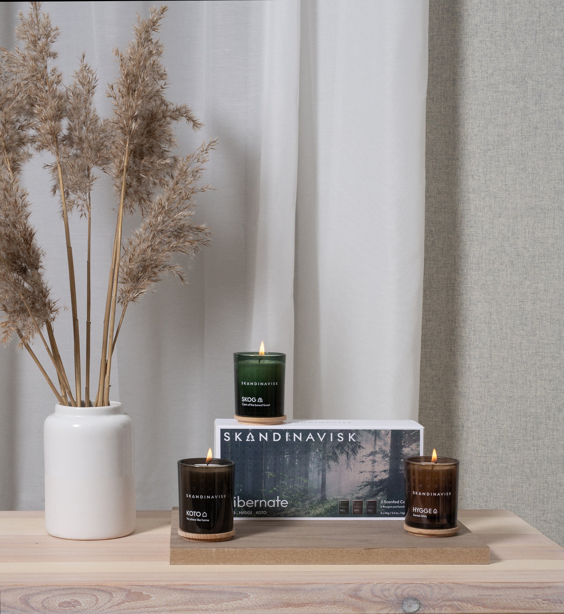Three lit mini scented candles from the Skandinavisk Giftset Scented Candles Hibernate—featuring SKOG, KOTO, and HYGGE fragrances—are arranged on a wooden shelf beside a white vase filled with dried grass. Behind the candles is a box labeled "Skandinavisk Hibernate." Sheer curtains hang softly in the background, creating an inviting atmosphere that makes this set a thoughtful gift or a cozy addition to any home.