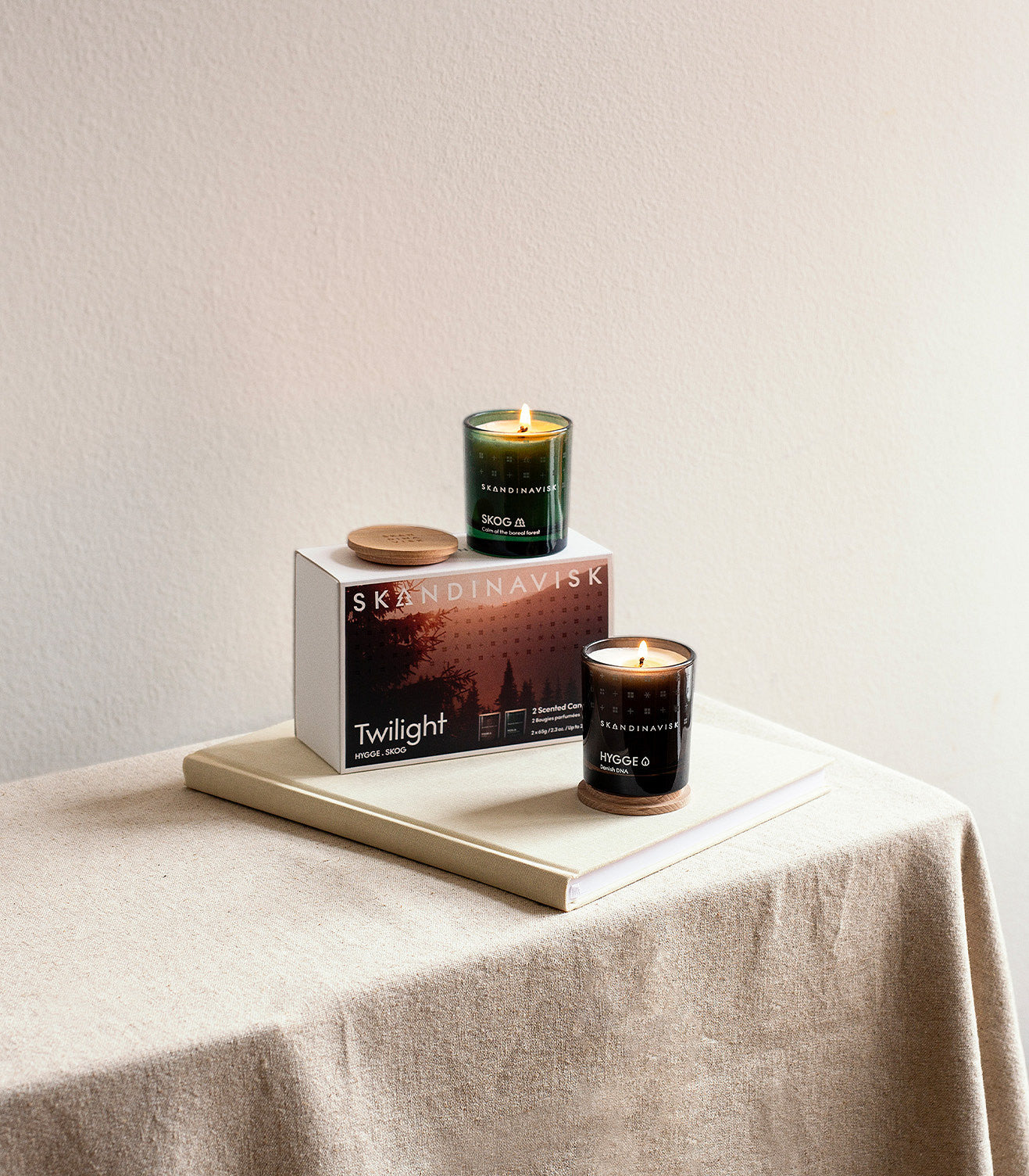 Two lit candles from the Skandinavisk Giftset Scented Candle Twilight with 2 Mini Candles Skog & Hygge are displayed in glass jars atop stacked books and a box labeled Twilight, resting on a beige tablecloth against a plain, light-colored wall. The minimalistic arrangement evokes the tranquil, cozy ambiance reminiscent of Scandinavian forest candles.