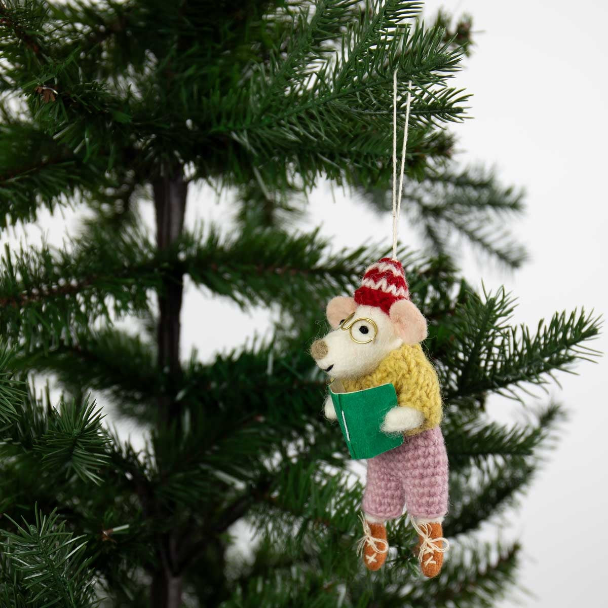 A Reading Teacher Christmas Ornament, Handmade in Wool, depicts a felted mouse in glasses, a patterned winter hat, a yellow sweater, and pink pants, holding a green book while hanging on a Christmas tree.